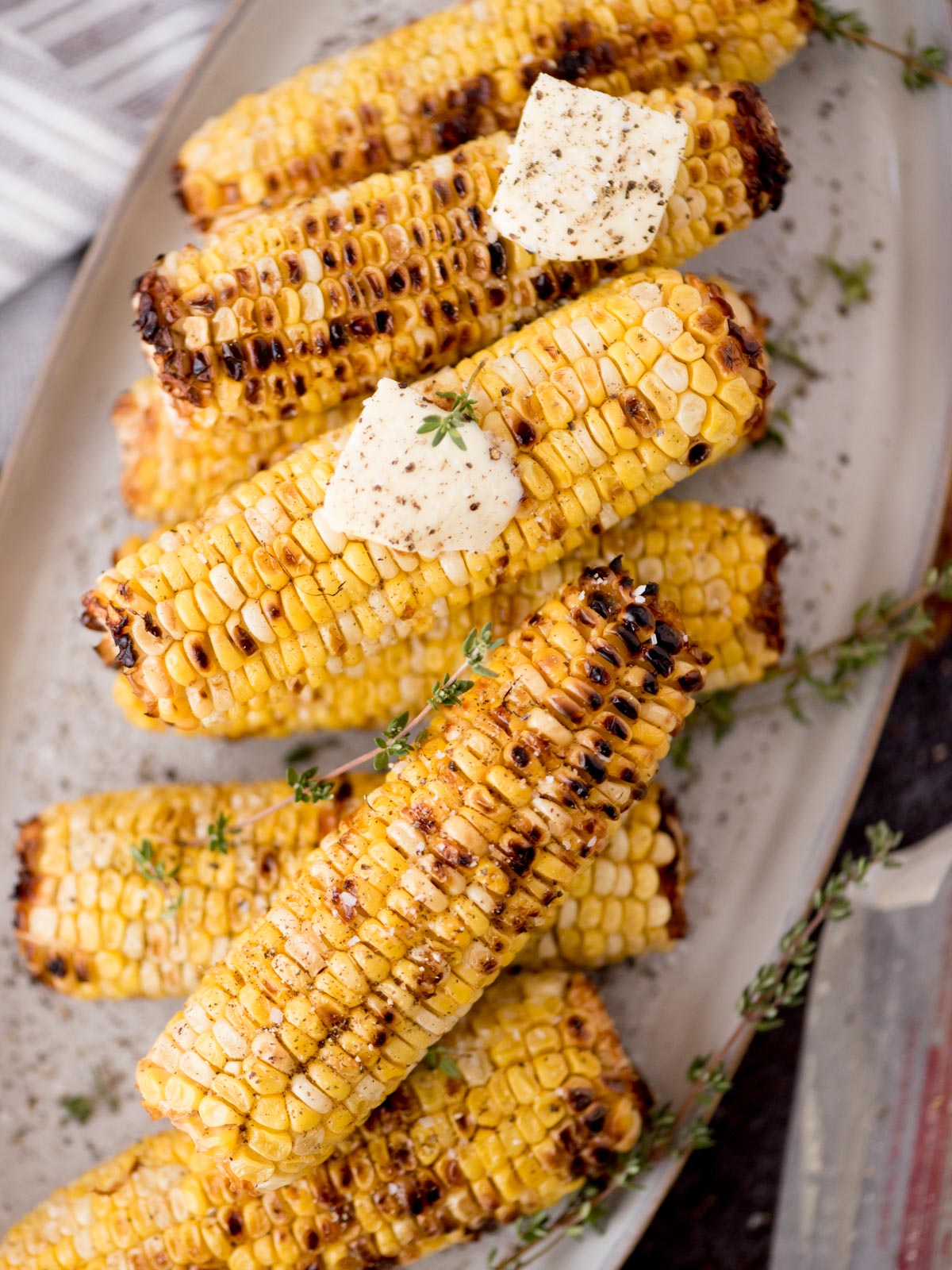 Grilled corn on the cob stacked on a plate, topped with melting butter, fresh thyme, and a sprinkle of black pepper.