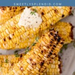 Grilled corn on the cob stacked on a plate, topped with melting butter and garnished with fresh herbs. Text overlay reads grilled corn and sweetlysplendid.com.