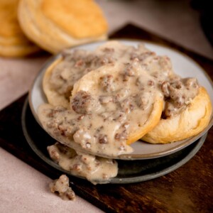 A plate of biscuits covered with creamy sausage gravy. The biscuits are fluffy and golden, with pieces of cooked sausage visible in the thick, peppered gravy. Another biscuit is in the background on the wooden surface.
