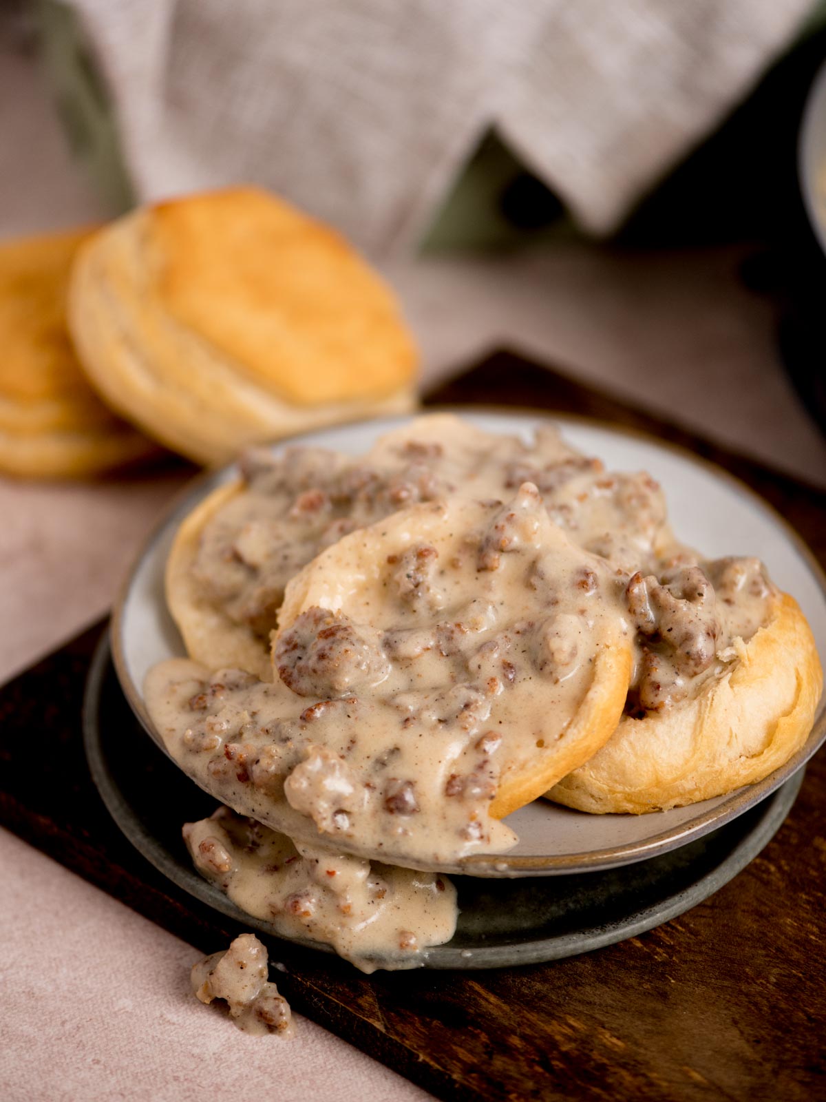 A plate of freshly baked biscuits topped with creamy sausage gravy. Additional biscuits are seen in the background on a wooden surface. The scene suggests a hearty, homemade breakfast setting.