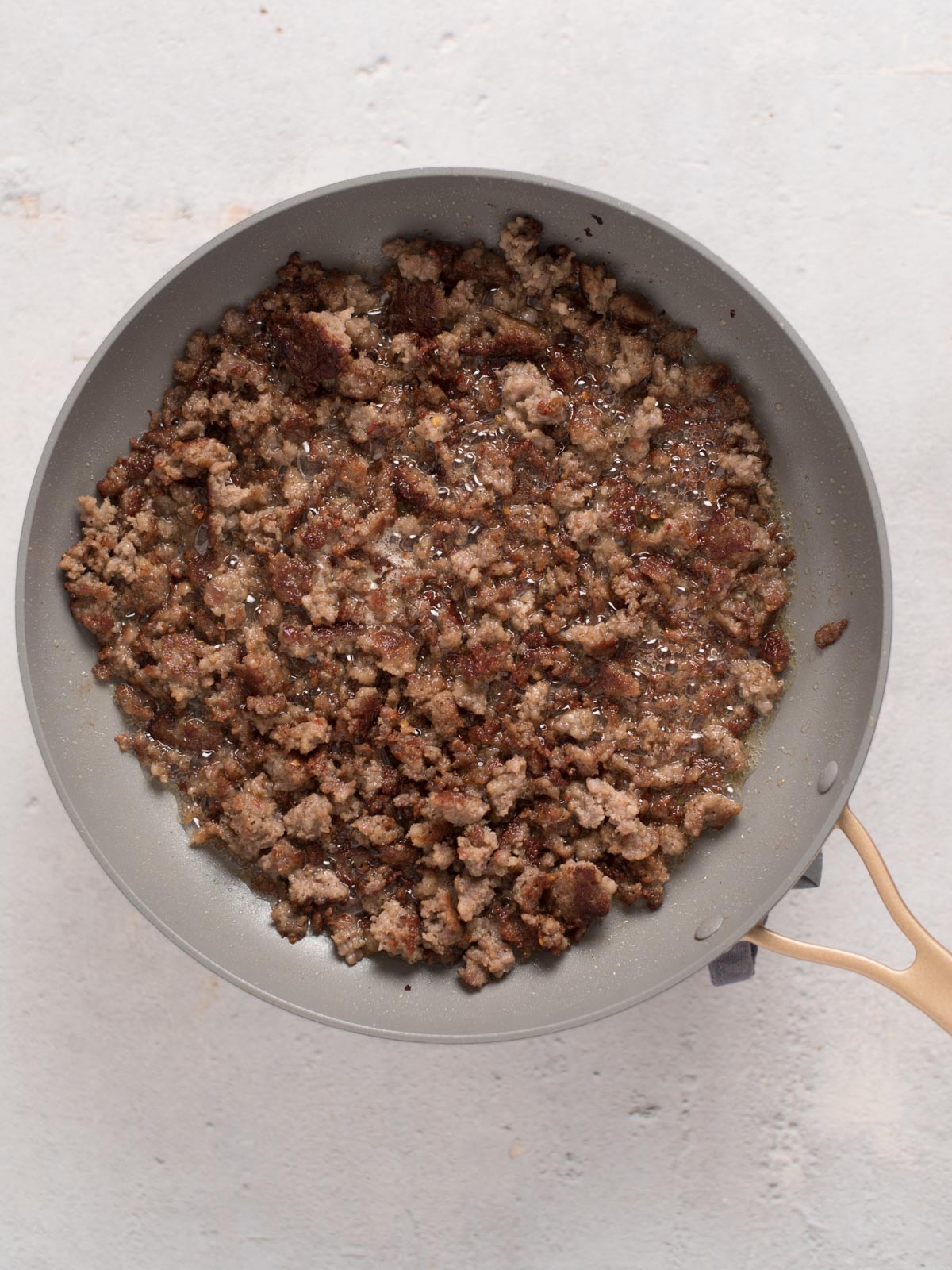 A pan containing browned and crumbled ground meat is placed on a light surface. The meat is cooked evenly, with a slightly crispy texture. The pan has a light gray interior and a wooden handle.