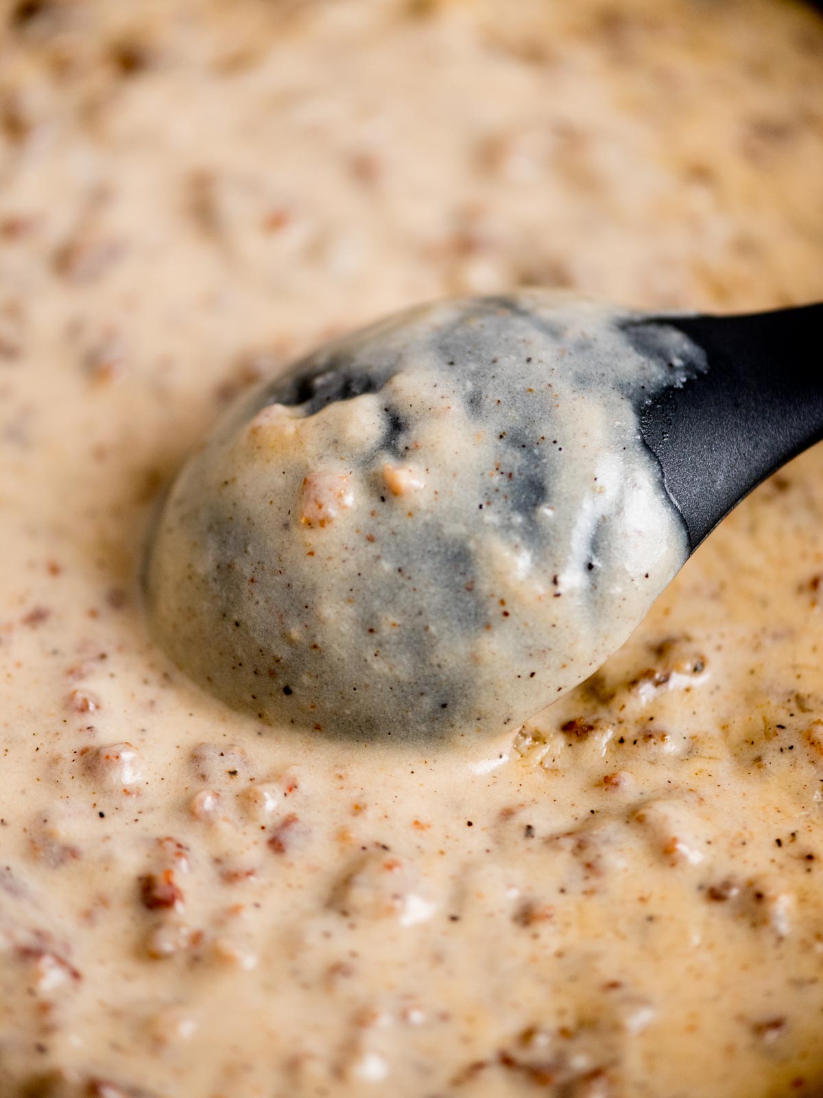 Close-up of a creamy gravy with ground sausage bits, being stirred by a black spoon. The gravy has a speckled appearance, indicating the presence of pepper or spices.