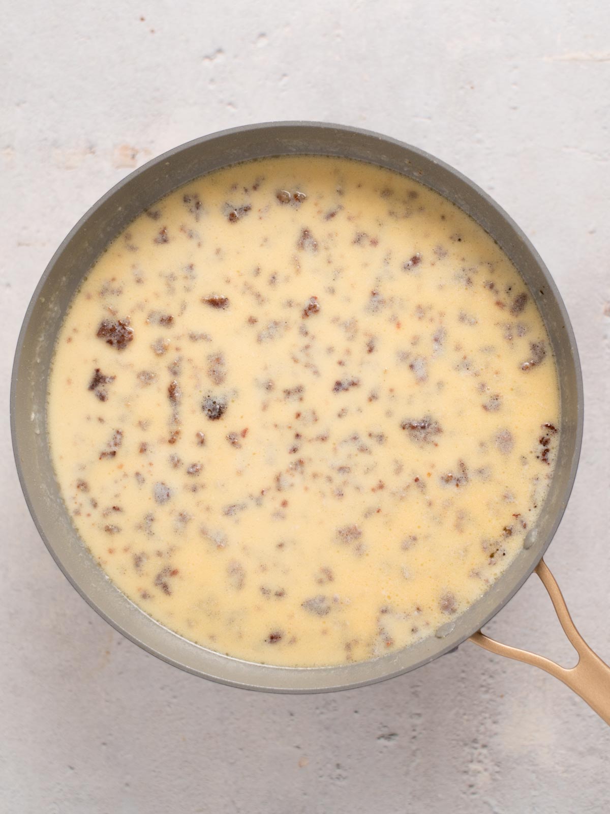 A pan filled with a creamy yellow mixture speckled with chunks of ground sausage. The mixture has a smooth, slightly thick texture and is set against a light gray background.