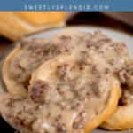 A plate of biscuits topped with sausage gravy. Text at the top reads Sausage Gravy with sweetlysplendid.com underneath. The plate is set on a wooden surface.