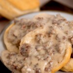 Biscuits smothered in creamy sausage gravy are stacked on a plate. Text at the top reads, creamy southern sausage gravy, with a website link below it. Another biscuit is partly visible in the background.