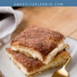 A plate holds two square slices of sopapilla cheesecake, their cinnamon sugar crust glistening invitingly. A gold fork rests nearby, ready to dive in. Sopapilla Cheesecake is inscribed above a website URL, while a napkin lies next to the delicious scene on the wooden table.