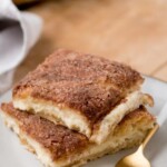 Two square slices of sopapilla cheesecake with a cinnamon sugar topping are elegantly stacked on a white plate. A gold fork rests beside them, ready to dive into the sweet delight. At the top, the text reads sopapilla cheesecake sweetlysplendid.com.