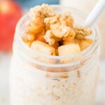 A glass jar filled with apple cinnamon overnight oats, topped with diced apples and granola, with a white spoon inside. A blurred red apple is in the background. The text reads apple cinnamon overnight oats.