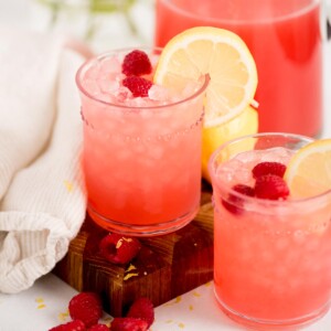 Two glasses of pink raspberry lemonade with ice, garnished with lemon slices and raspberries, sit on a wooden board next to a white cloth and a pitcher of the same drink. Fresh raspberries are scattered around.
