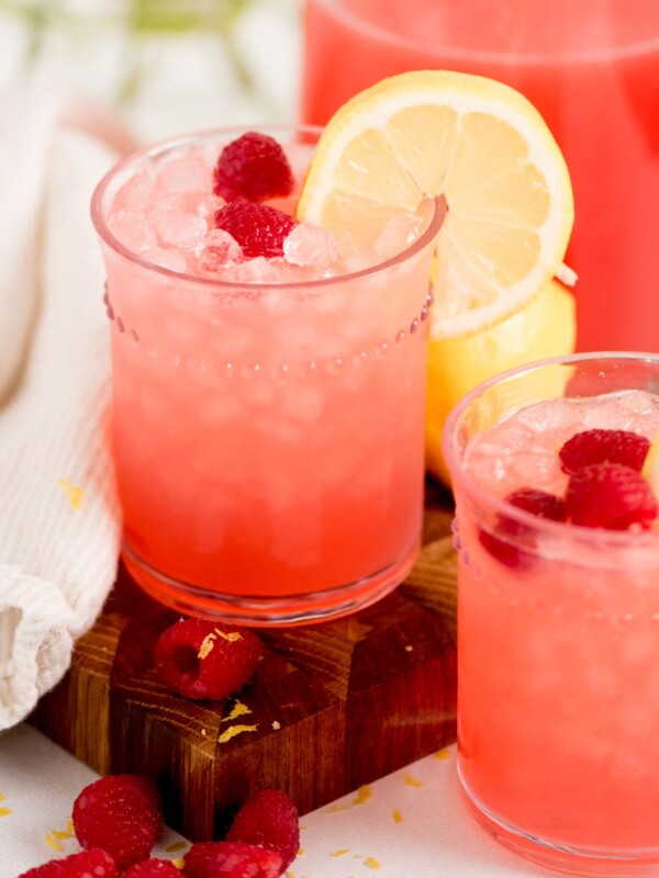 Two glasses of pink raspberry lemonade with ice, garnished with lemon slices and raspberries, sit on a wooden board next to a white cloth and a pitcher of the same drink. Fresh raspberries are scattered around.