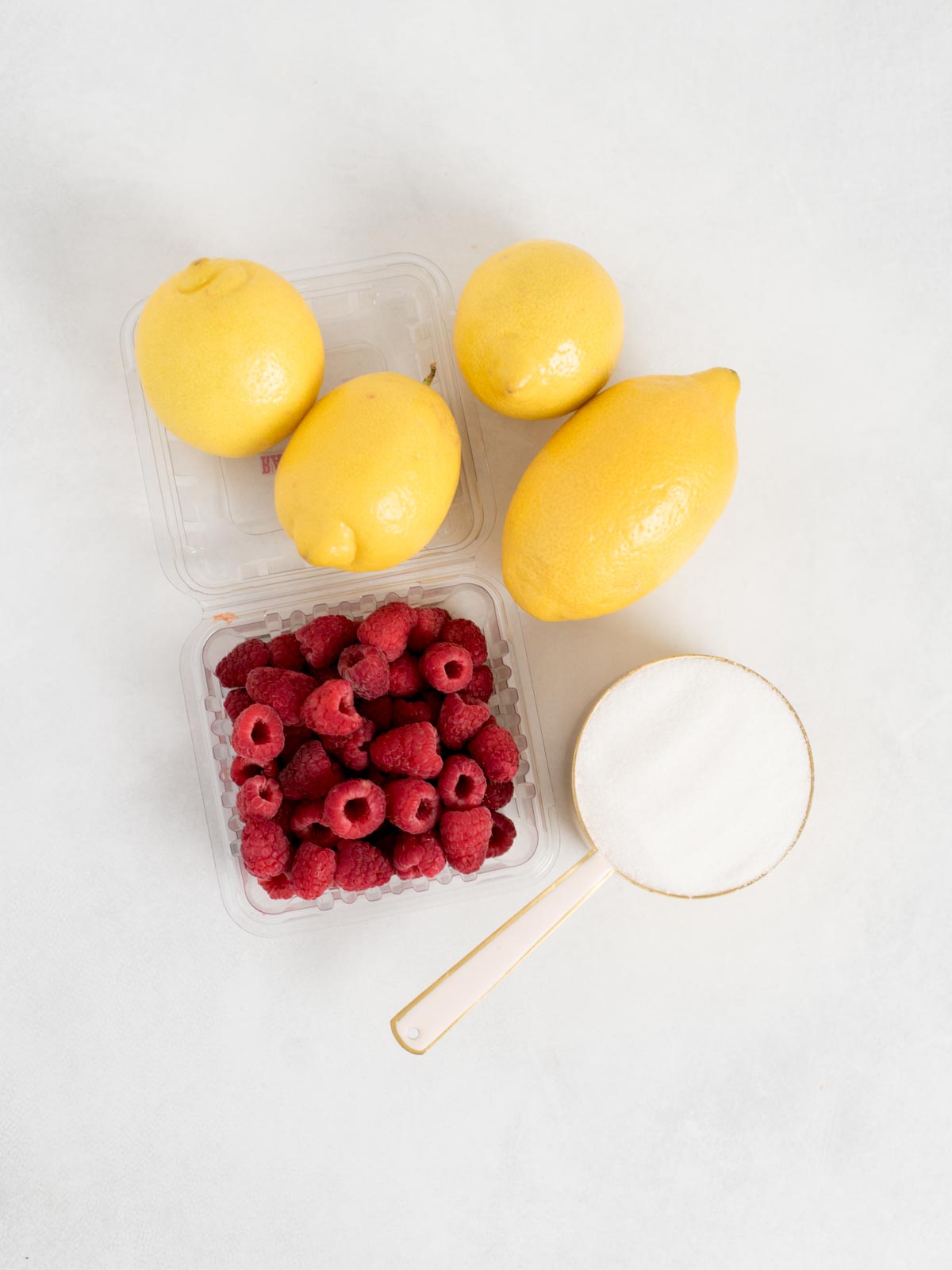Four lemons, a container of fresh raspberries, and a measuring cup filled with granulated sugar are arranged on a white surface.