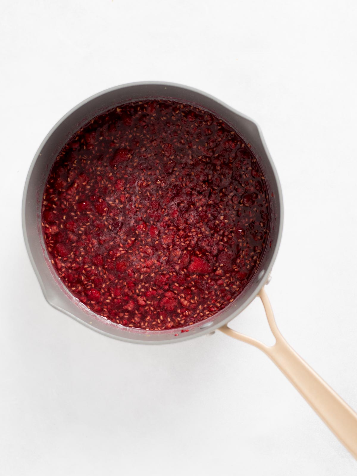 A gray saucepan with a beige handle filled with simmering raspberry sauce or jam, resting on a white surface.