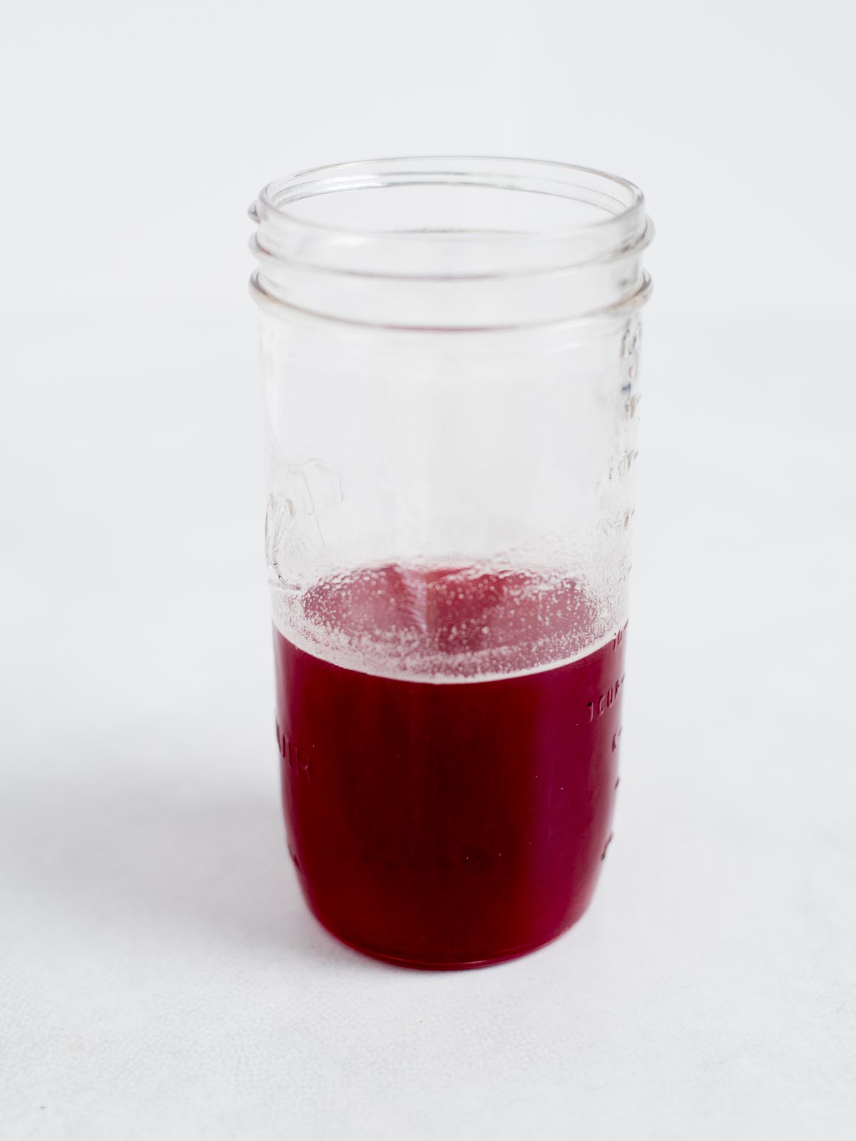 A clear glass jar containing a small amount of bright red liquid, placed on a white surface with a plain white background.