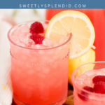 A glass of pink lemonade with ice, fresh raspberries, and a lemon slice garnish sits on a wooden board, surrounded by more raspberries and a lemon, with Pink Lemonade text above.