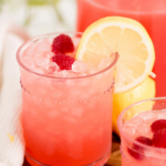 Three glasses of pink raspberry lemonade, garnished with lemon slices and raspberries, sit on a wooden board. The text above reads “fresh raspberry lemonade” and “sweetlysplendid.com.”.