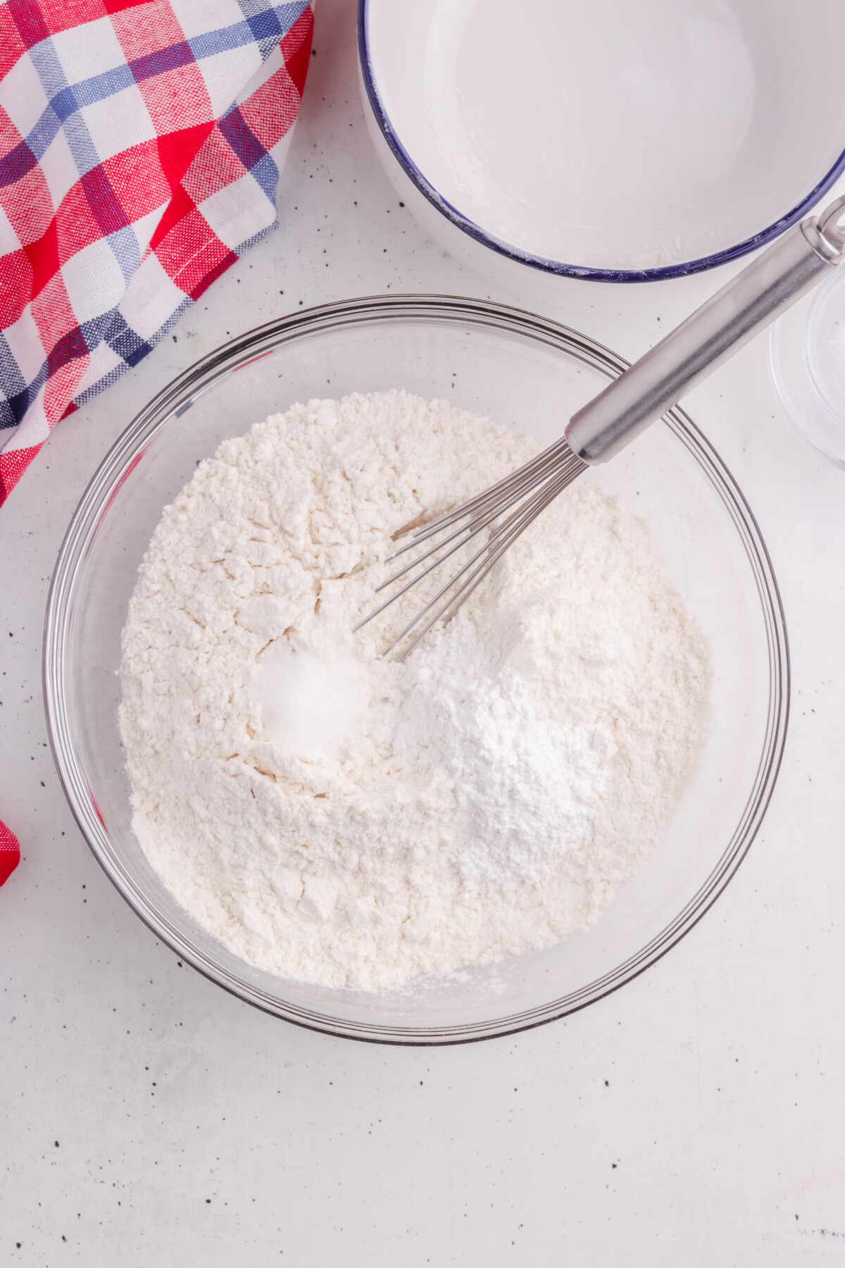 A glass bowl filled with flour and baking powder is being whisked. A red and blue checkered cloth and a white mixing bowl are nearby on a white countertop.