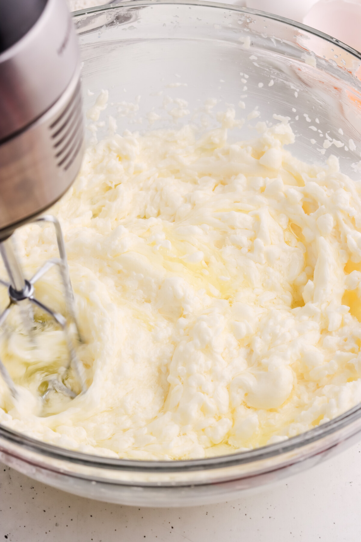 A hand mixer blending a creamy, pale mixture in a glass bowl, likely butter and sugar being whipped together on a white countertop.
