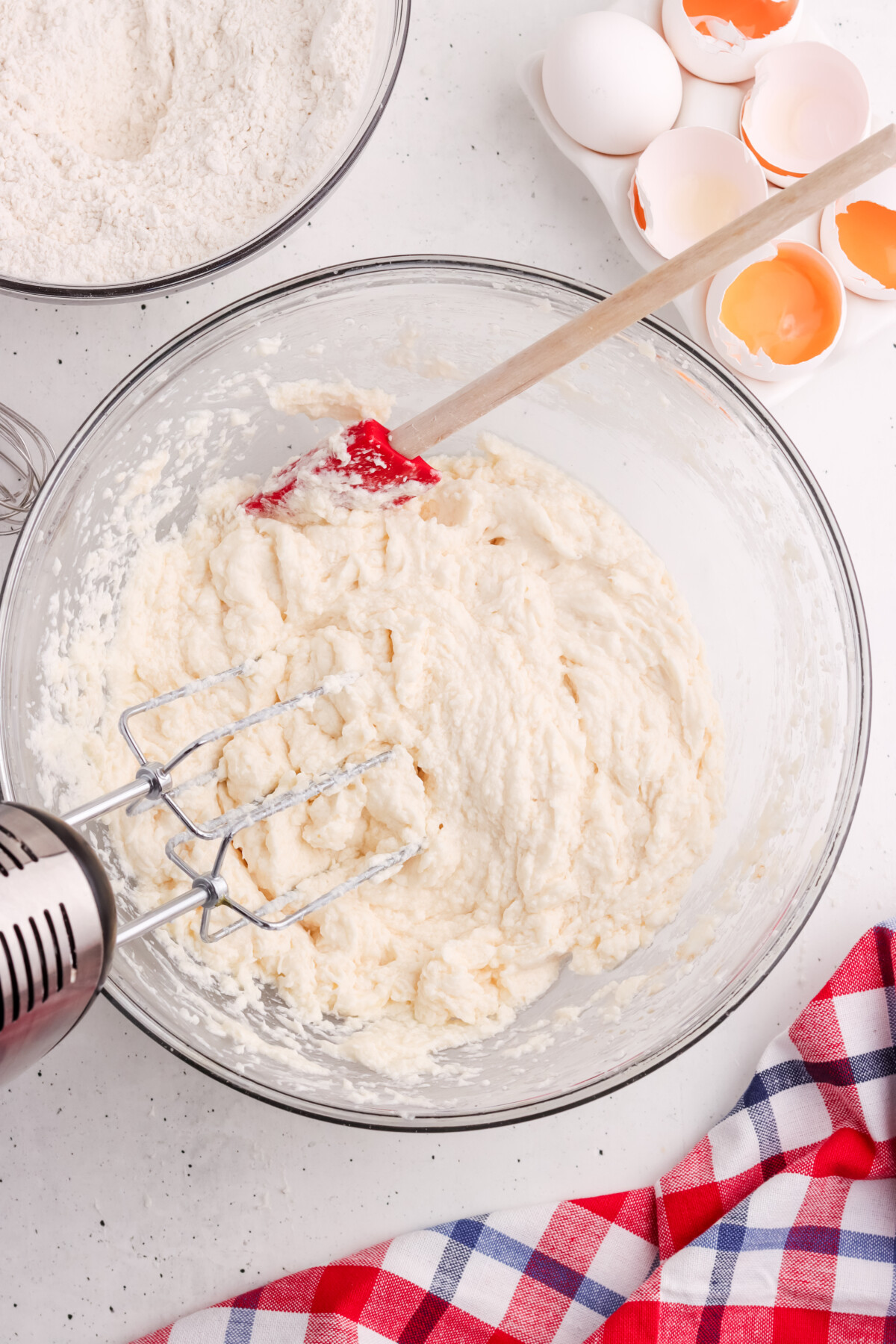 A glass bowl with cake batter being mixed by a hand mixer and red spatula, next to cracked eggshells, a bowl of flour, and a red plaid cloth on a white countertop.
