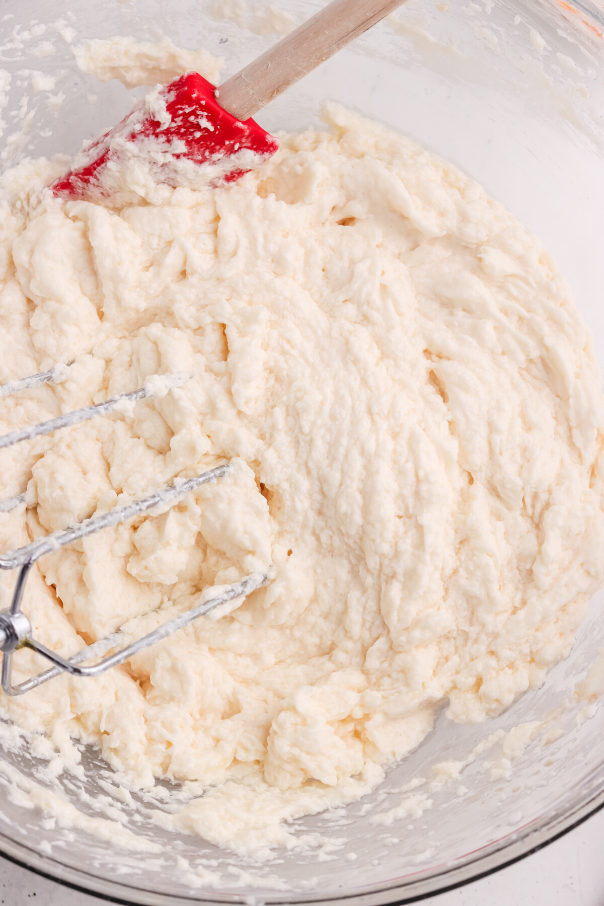A glass bowl filled with thick, creamy cake batter. A red spatula and metal hand mixer beaters rest in the batter, showing the mixing process in progress.