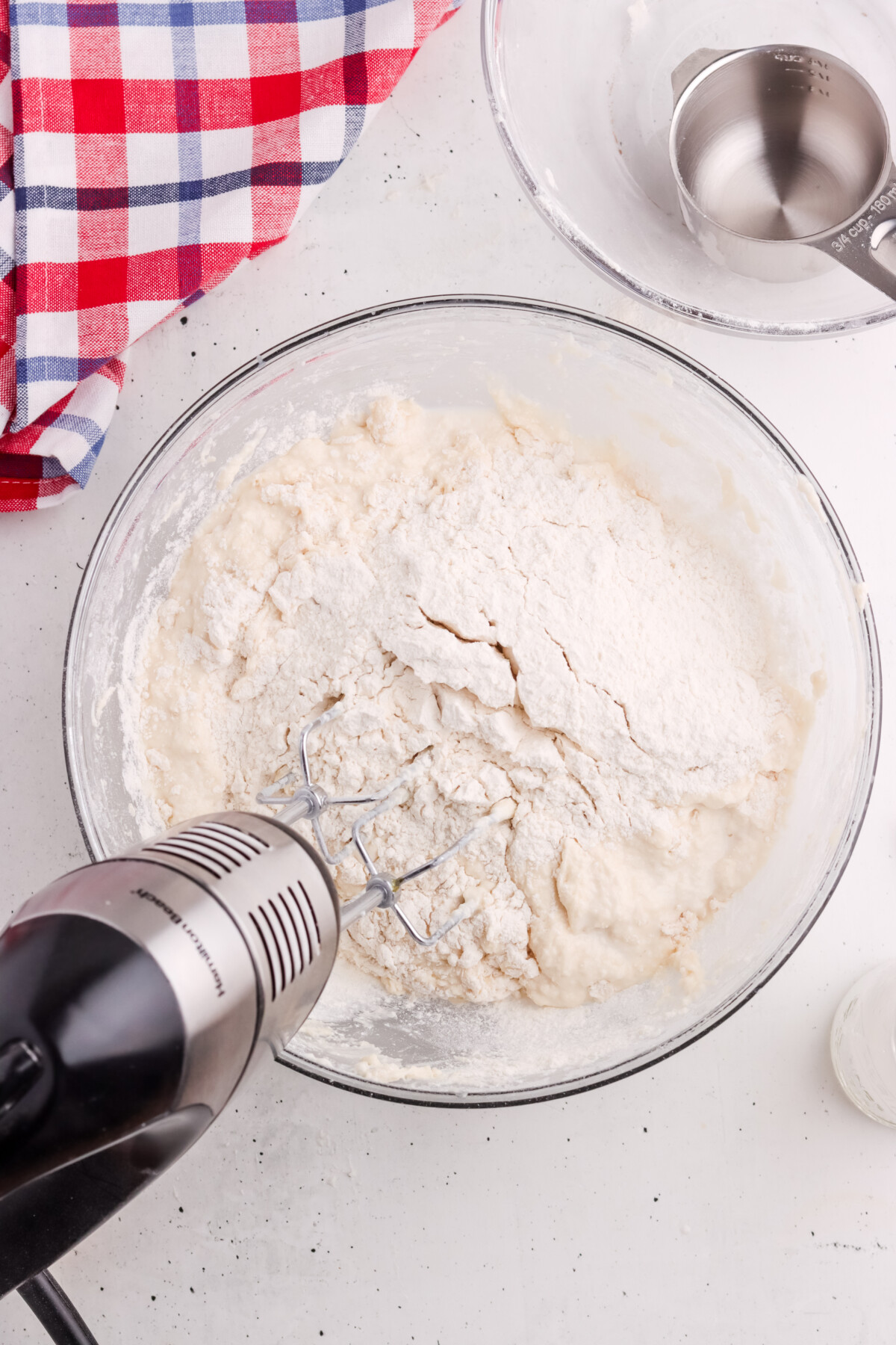 A glass bowl with flour and dough being mixed by an electric hand mixer sits on a white surface near a red, white, and blue checkered cloth and a metal measuring cup.