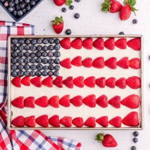 A rectangular cake decorated as an American flag with blueberries in the corner and rows of halved strawberries on white frosting. Surrounding the cake are fresh strawberries, blueberries, and a red, white, and blue cloth.