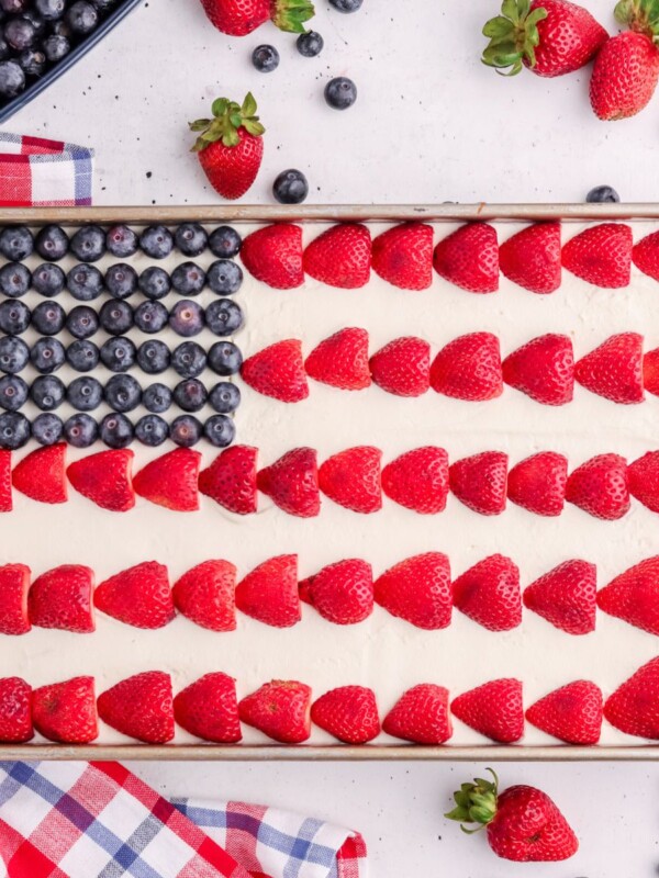 A rectangular cake decorated as an American flag with blueberries in the corner and rows of halved strawberries on white frosting. Surrounding the cake are fresh strawberries, blueberries, and a red, white, and blue cloth.