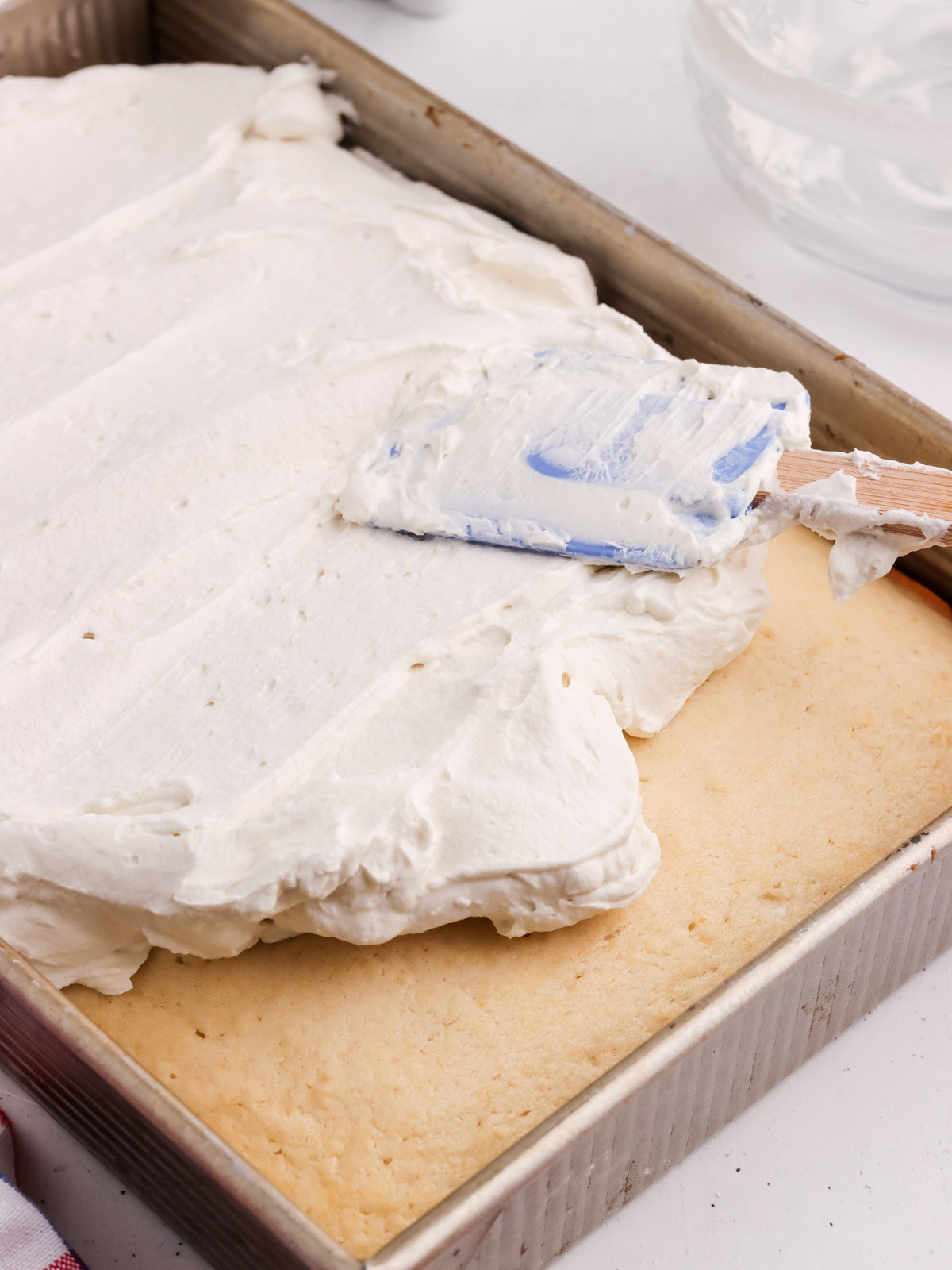 A metal baking pan holds an American Flag Cake, as a thick layer of white icing is smoothed over the rectangular cake with a blue and white spatula.