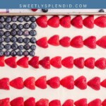 A sheet cake decorated as an American flag with blueberries for stars and rows of halved strawberries for stripes. A checkered napkin and fresh strawberries are on the table beside the cake.