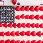 Rectangular cake decorated as an American flag with blueberries in the top left corner for stars and rows of strawberries on white frosting for stripes. A checkered cloth and fresh berries are nearby.