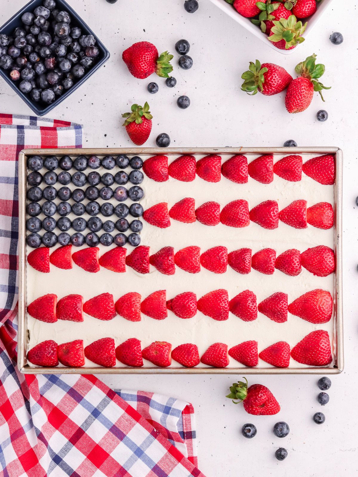A sheet cake decorated like the American flag with blueberries in the top left and rows of sliced strawberries on white frosting. Fresh strawberries and blueberries are scattered around on a plaid napkin.