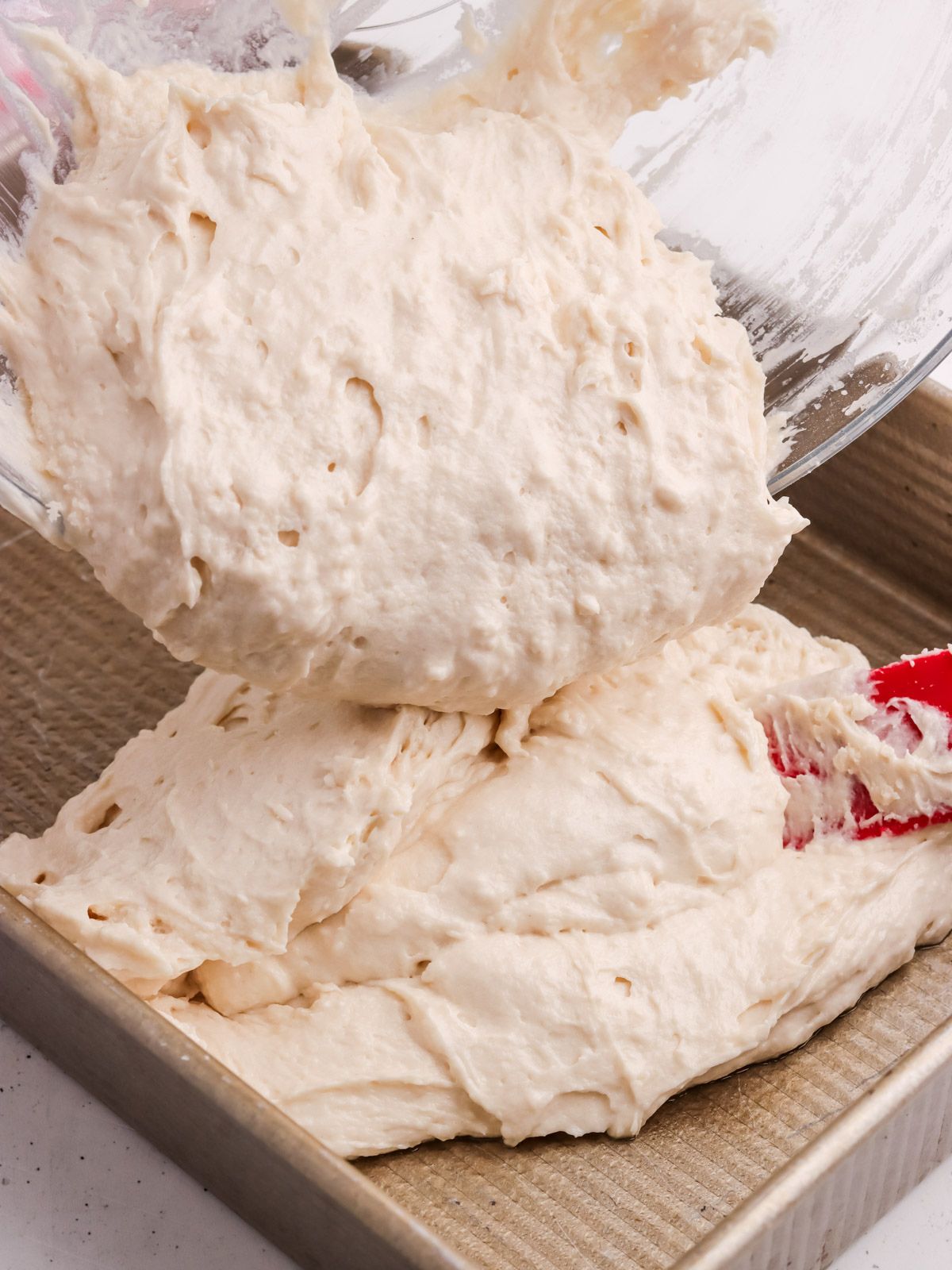 Thick bread dough is being poured from a glass bowl into a square metal baking pan with the help of a red spatula.