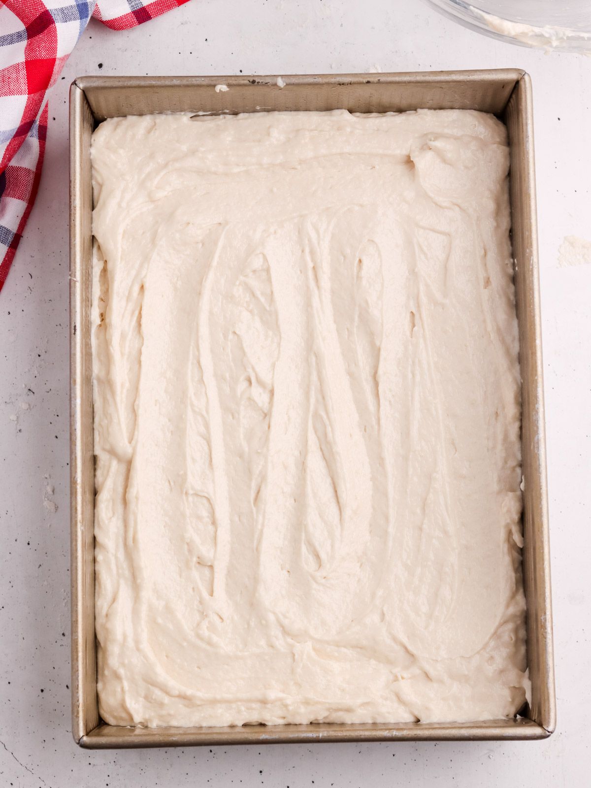 A rectangular metal baking pan filled with smooth, creamy cake batter, spread evenly across the surface. A red, white, and blue checkered cloth is partially visible in the corner.