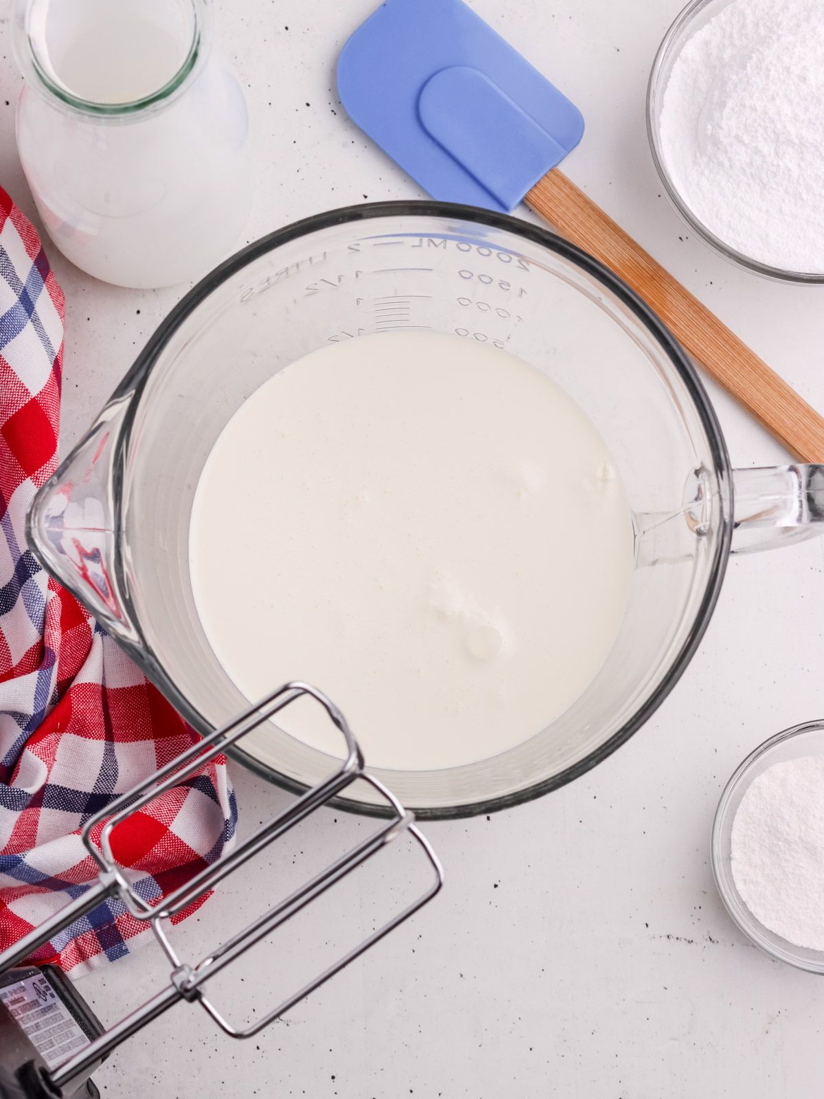 A glass mixing bowl filled with milk or cream sits on a white surface, surrounded by a checkered cloth, a bottle of milk, a blue spatula, a bowl of sugar, and a hand mixer.