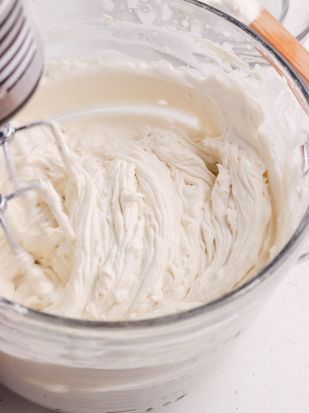 A close-up of thick, creamy frosting being mixed in a clear glass bowl with an electric hand mixer, creating soft peaks and swirls in the frosting.