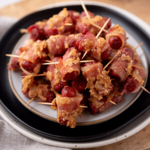 A plate of bacon-wrapped cocktail sausages held together with toothpicks, with a golden-brown, crispy coating on the bacon, arranged in a pile on a white and black plate.