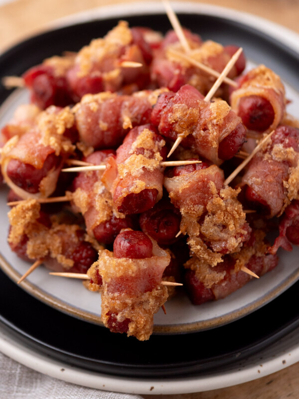 A plate of bacon-wrapped cocktail sausages held together with toothpicks, with a golden-brown, crispy coating on the bacon, arranged in a pile on a white and black plate.