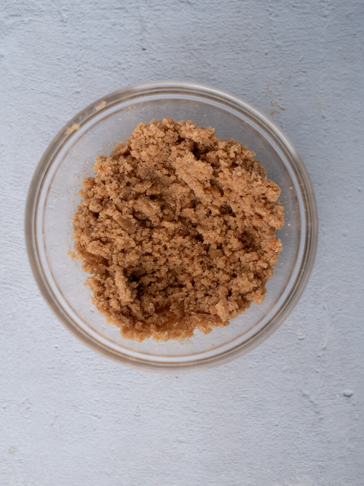 A glass bowl filled with brown sugar sits on a light gray textured surface, viewed from above.