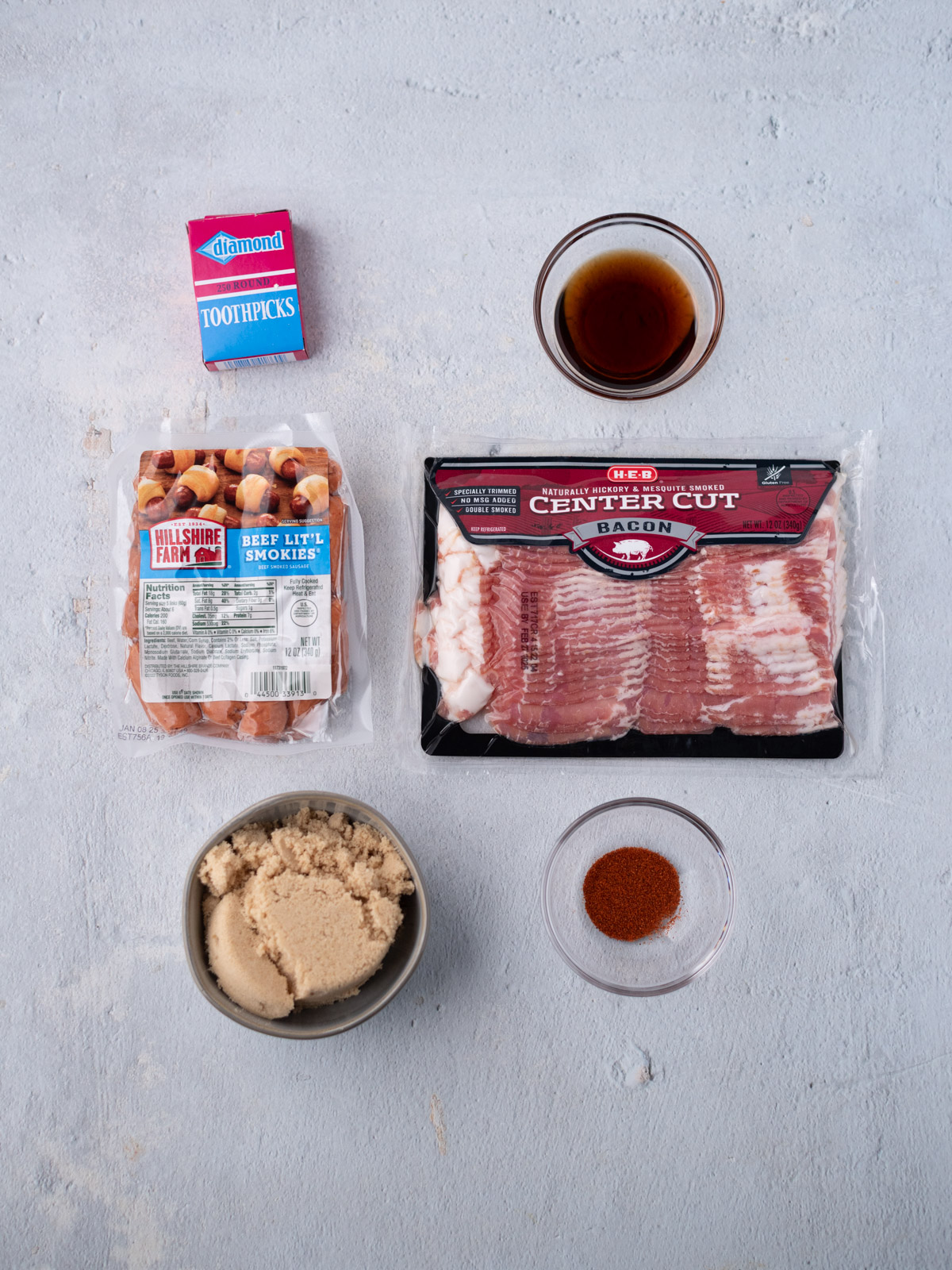 Five ingredients and toothpicks arranged on a light surface: a pack of beef little smokies, center cut bacon, brown sugar in a bowl, cayenne in a small bowl, a bowl of maple syrup, and a box of toothpicks.