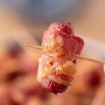 A close-up of a small sausage wrapped in crispy bacon, held on a toothpick. Text at the top reads brown sugar bacon smokies with the website sweetlysplendid.com underneath.