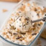 A metal ice cream scooper lifting a scoop of creamy ice cream with chocolate chips and crushed cookies from a glass container. Crumbled cookies are sprinkled on top.