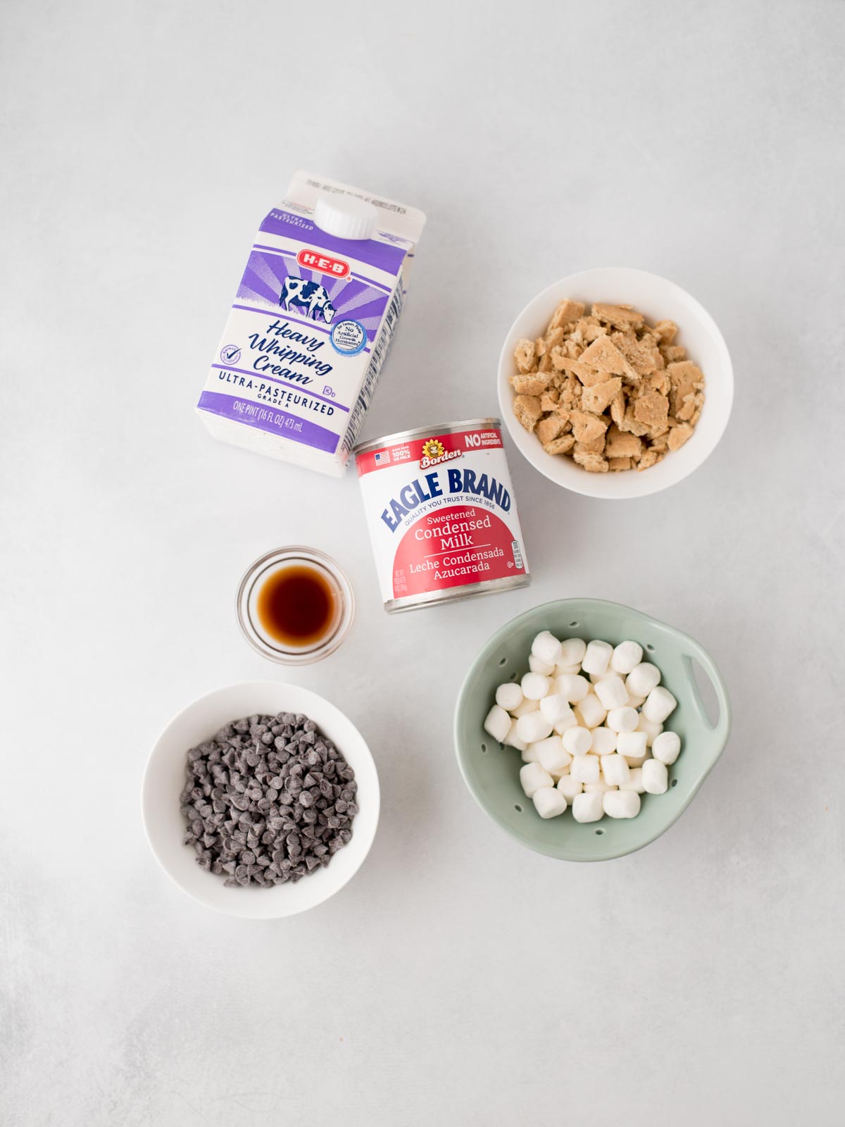 Five ingredients on a light surface: a carton of heavy whipping cream, a bowl of brown sugar, a can of condensed milk, a bowl of mini chocolate chips, a bowl of mini marshmallows, and a small cup of vanilla extract.