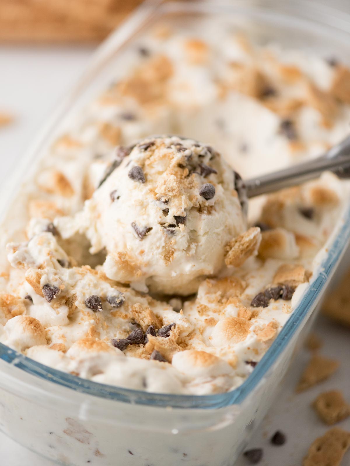 Close-up of a glass dish filled with creamy ice cream mixed with chocolate chips and crushed graham crackers, with a metal scoop lifting a portion.