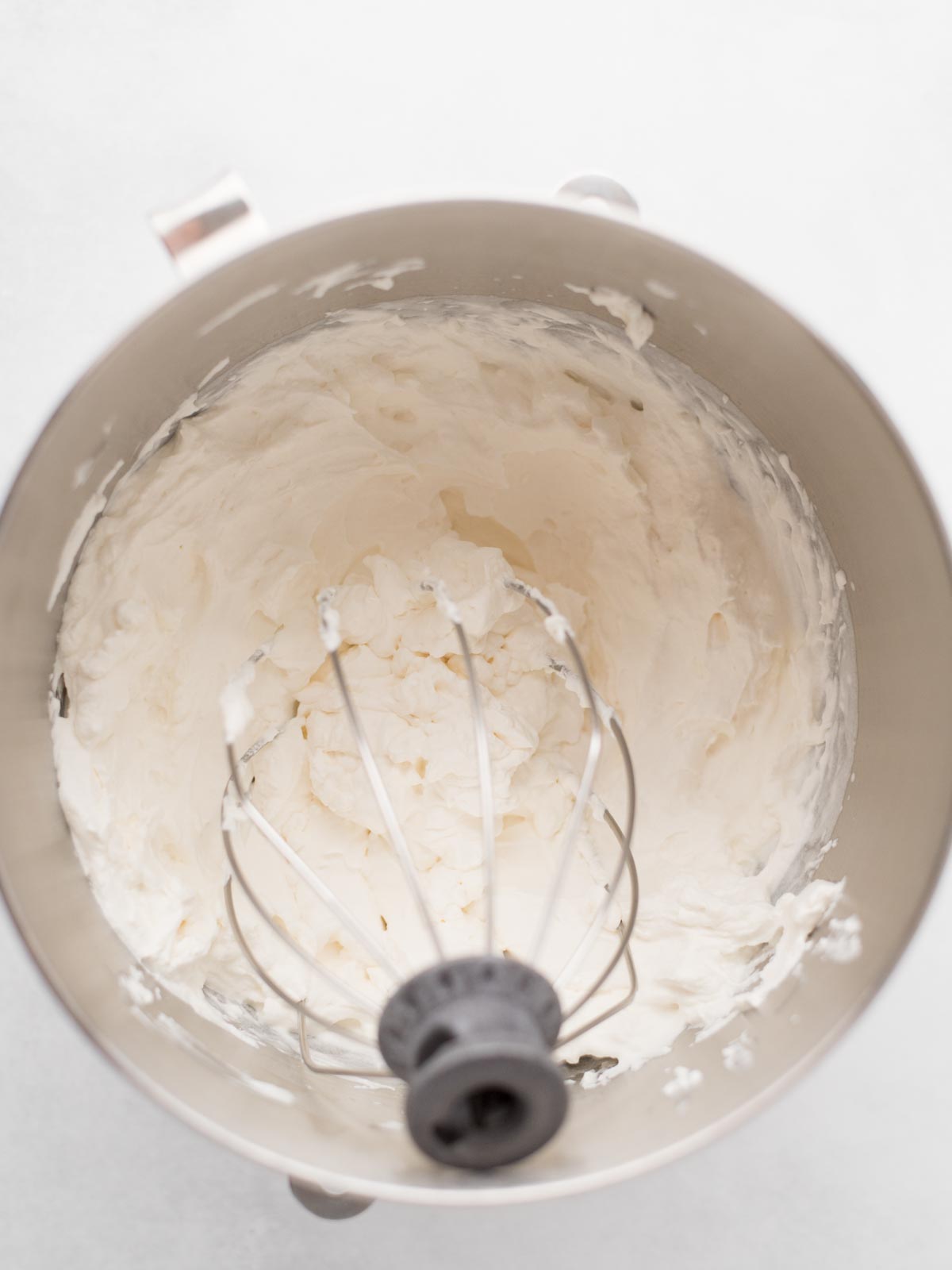 A metal mixing bowl with a whisk attachment contains freshly whipped cream, with soft peaks visible and some cream clinging to the sides of the bowl. The background is a light, neutral color.