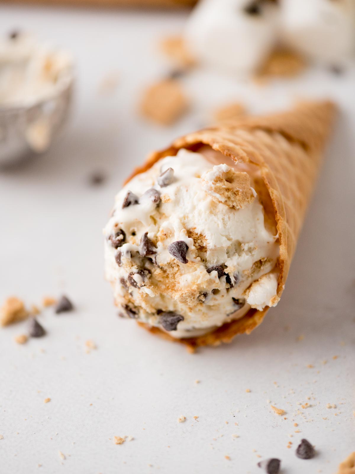 A waffle cone filled with a scoop of ice cream containing chocolate chips and cookie crumbs, lying on its side with some chocolate chips and crumbs scattered nearby on a light surface.
