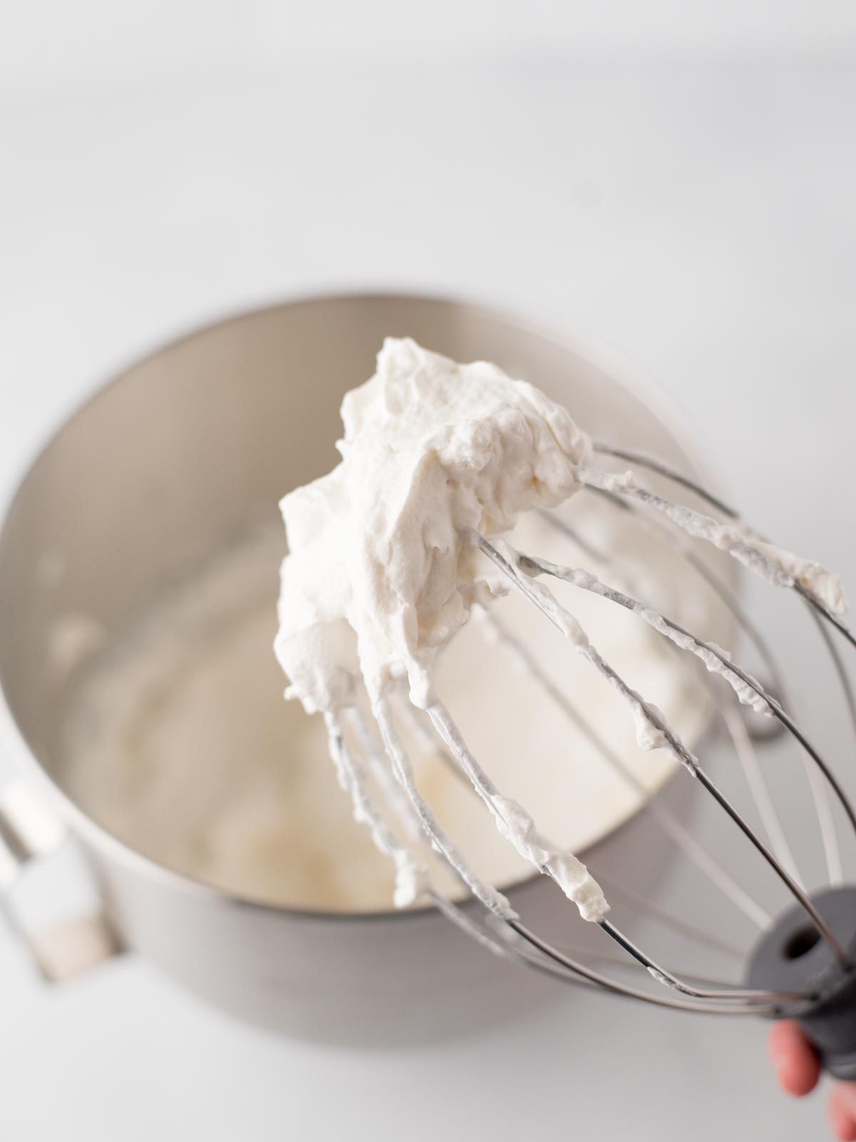 A close-up of a metal whisk covered in freshly whipped cream, held above a mixing bowl filled with more whipped cream. The background is softly blurred.