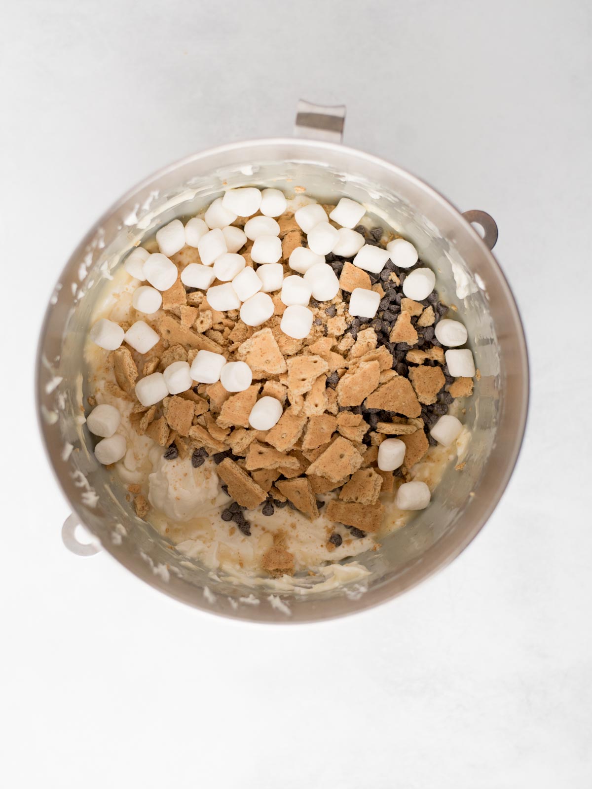 A mixing bowl containing mini marshmallows, chocolate chips, and broken graham crackers on top of a creamy mixture, viewed from above.