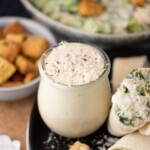 A small glass jar filled with creamy Caesar salad dressing sits on a plate next to a wrap and croutons, with a bowl of salad and a dish of croutons in the background.