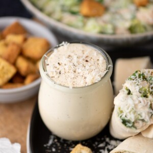 A small glass jar filled with creamy Caesar salad dressing sits on a plate next to a wrap and croutons, with a bowl of salad and a dish of croutons in the background.