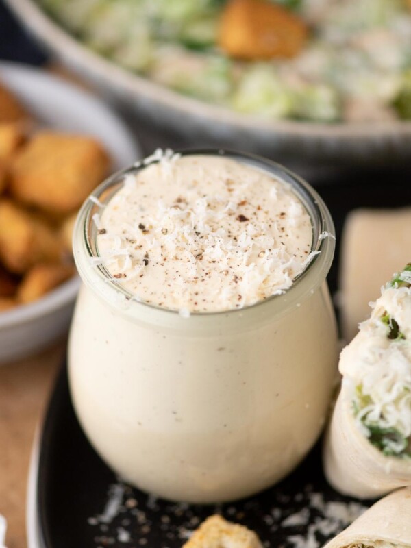 A small glass jar filled with creamy Caesar salad dressing sits on a plate next to a wrap and croutons, with a bowl of salad and a dish of croutons in the background.