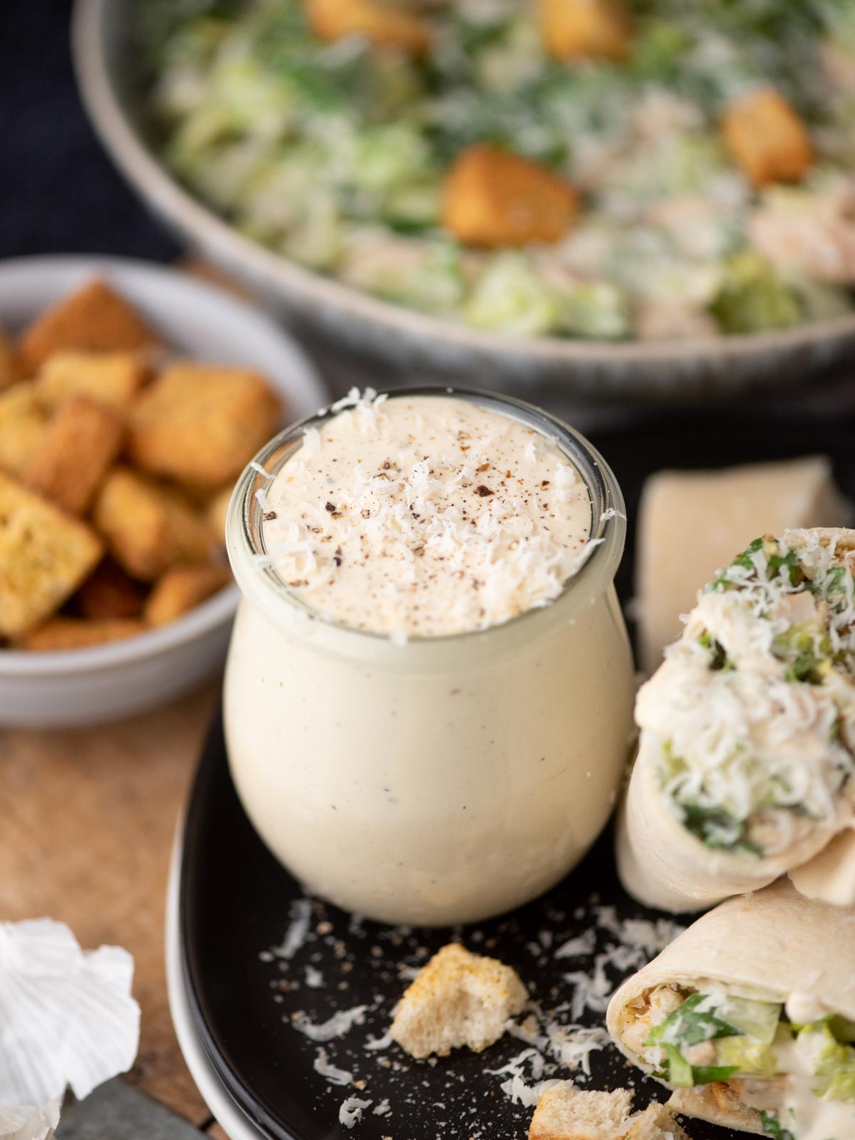 A jar of creamy Caesar salad dressing is surrounded by croutons, a wrap filled with salad, and a bowl of Caesar salad in the background. Grated cheese is sprinkled on top of the dressing and the wrap.